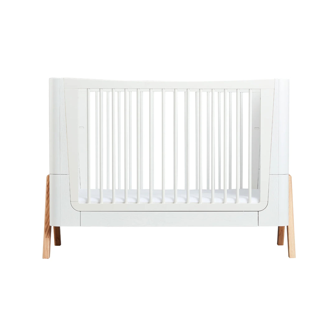 Front view of the Gaia Baby Hera Cot in Natural Scandi White, showcasing its modern white frame, natural wood legs, and safe slatted sides; premium eco-friendly Scandinavian-style cot bed for contemporary nursery design.