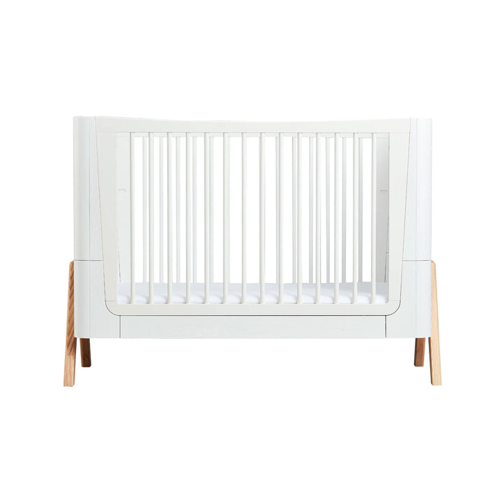 Front view of the Gaia Baby Hera Cot in Natural Scandi White, showcasing its modern white frame, natural wood legs, and safe slatted sides; premium eco-friendly Scandinavian-style cot bed for contemporary nursery design.
