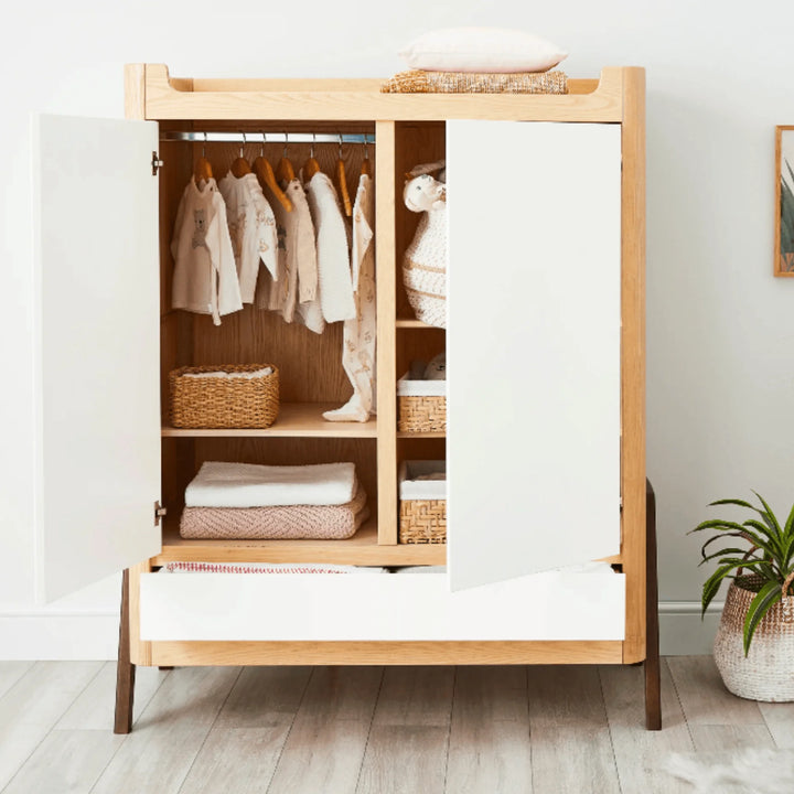 Gaia Baby Hera wardrobe in Natural Ash and Walnut shown open in a styled nursery, displaying hanging baby clothes, woven baskets and folded blankets on adjustable shelves with bottom drawer extended.
