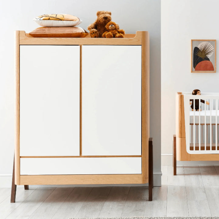 “Gaia Baby Hera wardrobe in Natural Ash and Walnut styled in a modern nursery, featuring white doors, solid ash frame, walnut legs and decorative accessories beside matching cot.”