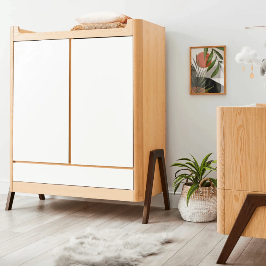 Gaia Baby Hera wardrobe in Natural Ash and Walnut styled in a modern Scandinavian nursery, featuring white doors, solid ash frame, walnut legs and coordinating wooden nursery furniture.