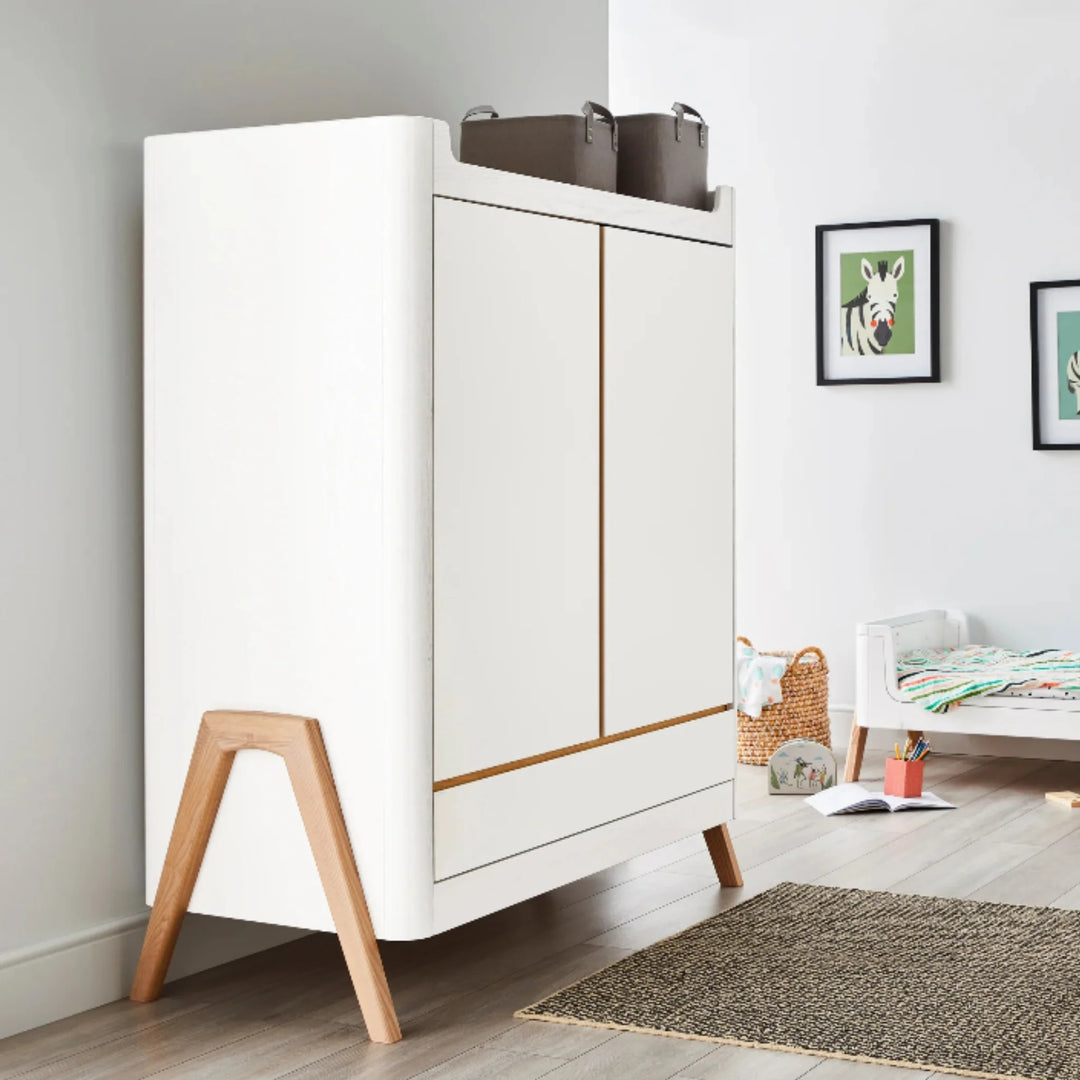 Gaia Baby Hera wardrobe in Scandi white and natural wood styled in a modern nursery, featuring angled wooden legs, storage baskets and children’s wall art for a Scandinavian design look
