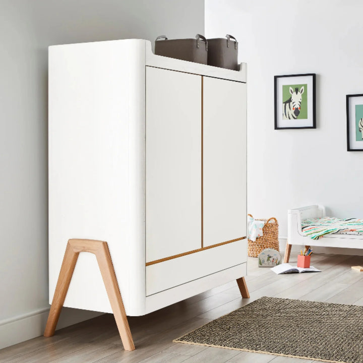 Gaia Baby Hera wardrobe in Scandi white and natural wood styled in a modern nursery, featuring angled wooden legs, storage baskets and children’s wall art for a Scandinavian design look