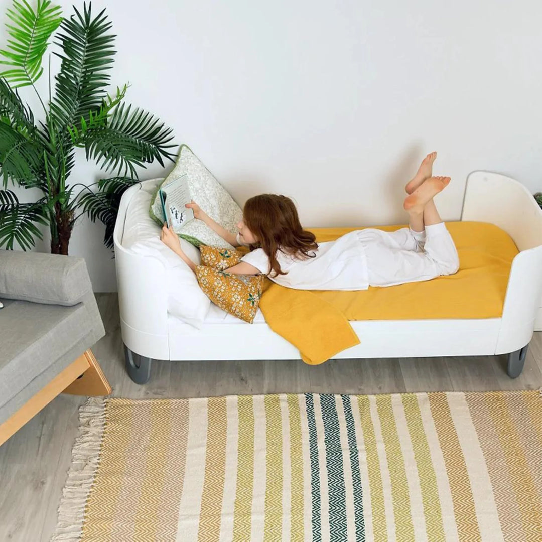 Child reading on Gaia Baby Serena Junior Bed in all-white design with extended cot bed conversion, styled with mustard blanket and decorative cushions in a modern kids’ bedroom.