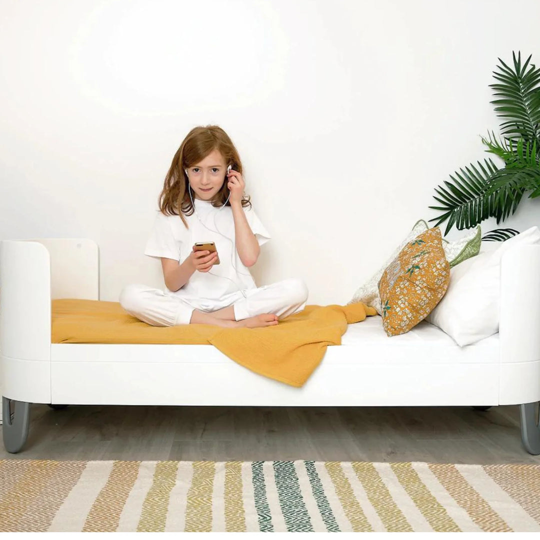 Child sitting on Gaia Baby Serena Junior Bed with natural legs and white frame, showcasing extended cot bed conversion in modern kids’ bedroom with mustard blanket and decorative cushions.