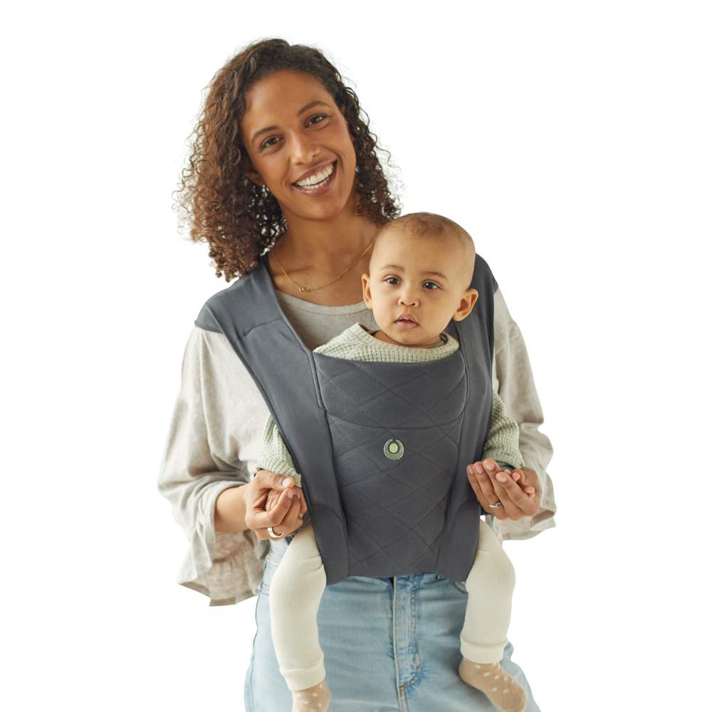 This image shows the Gaia Baby Soft Newborn+ Baby Carrier on a Mum who is smiling at the camera with her child in the world-facing position of the carrier, looking around inquizitively