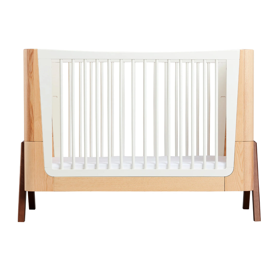 Gaia Baby Hera cot in ash and walnut, front-on view showcasing natural wood panels, white slatted sides, signature curved Scandinavian design and solid walnut A-frame legs — premium sustainable nursery furniture.