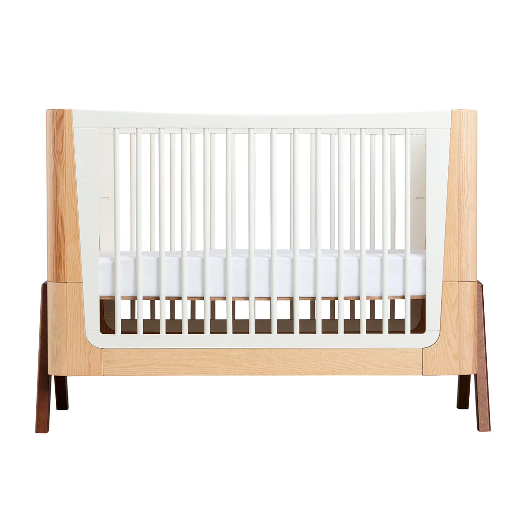 Gaia Baby Hera cot in ash and walnut, front view showing natural wood panels, white slatted sides, curved Scandinavian design and solid walnut A-frame legs — premium sustainable nursery furniture