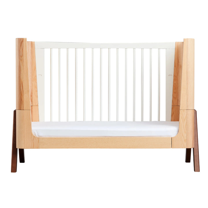 Gaia Baby Hera cot in ash and walnut, shown in toddler bed mode with natural wood panels, white slatted sides and solid walnut A-frame legs — premium Scandinavian sustainable nursery furniture.