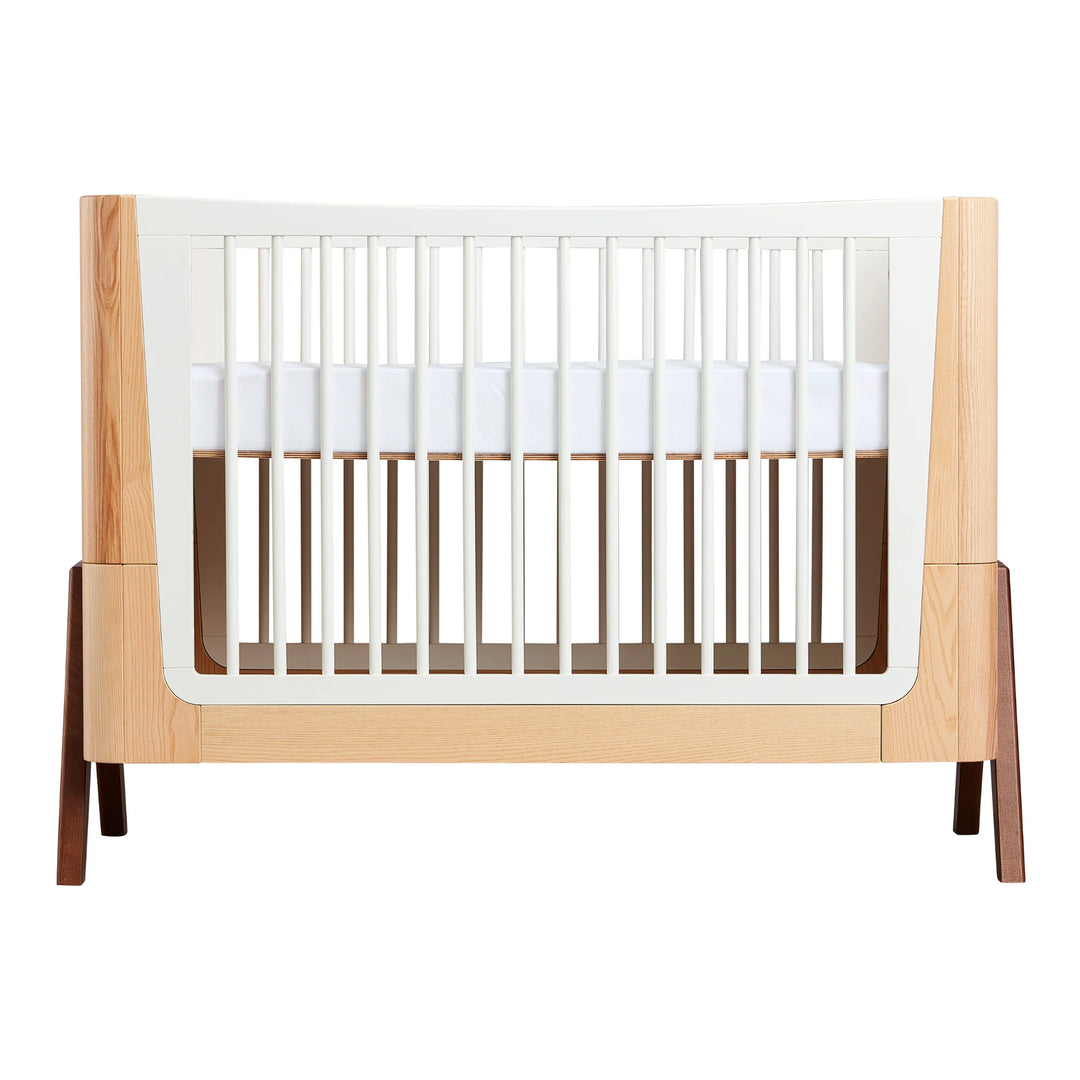 Gaia Baby Hera cot in ash and walnut, front view with mattress raised to newborn height, featuring natural wood panels, white slatted sides, curved Scandinavian styling and solid walnut A-frame legs — premium sustainable nursery cot.