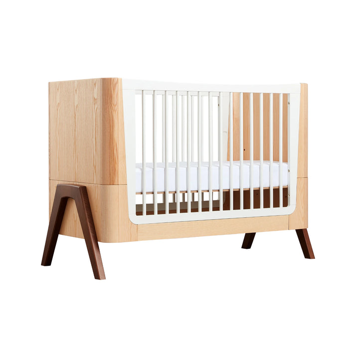 Gaia Baby Hera cot in ash and walnut, angled view showcasing natural wood panels, white slatted sides, curved Scandinavian design and solid walnut A-frame legs — premium sustainable nursery cot.