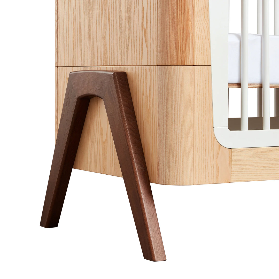 Close-up of Gaia Baby Hera cot in ash and walnut showing solid walnut A-frame legs, smooth curved wood detailing and premium Scandinavian sustainable nursery furniture craftsmanship.