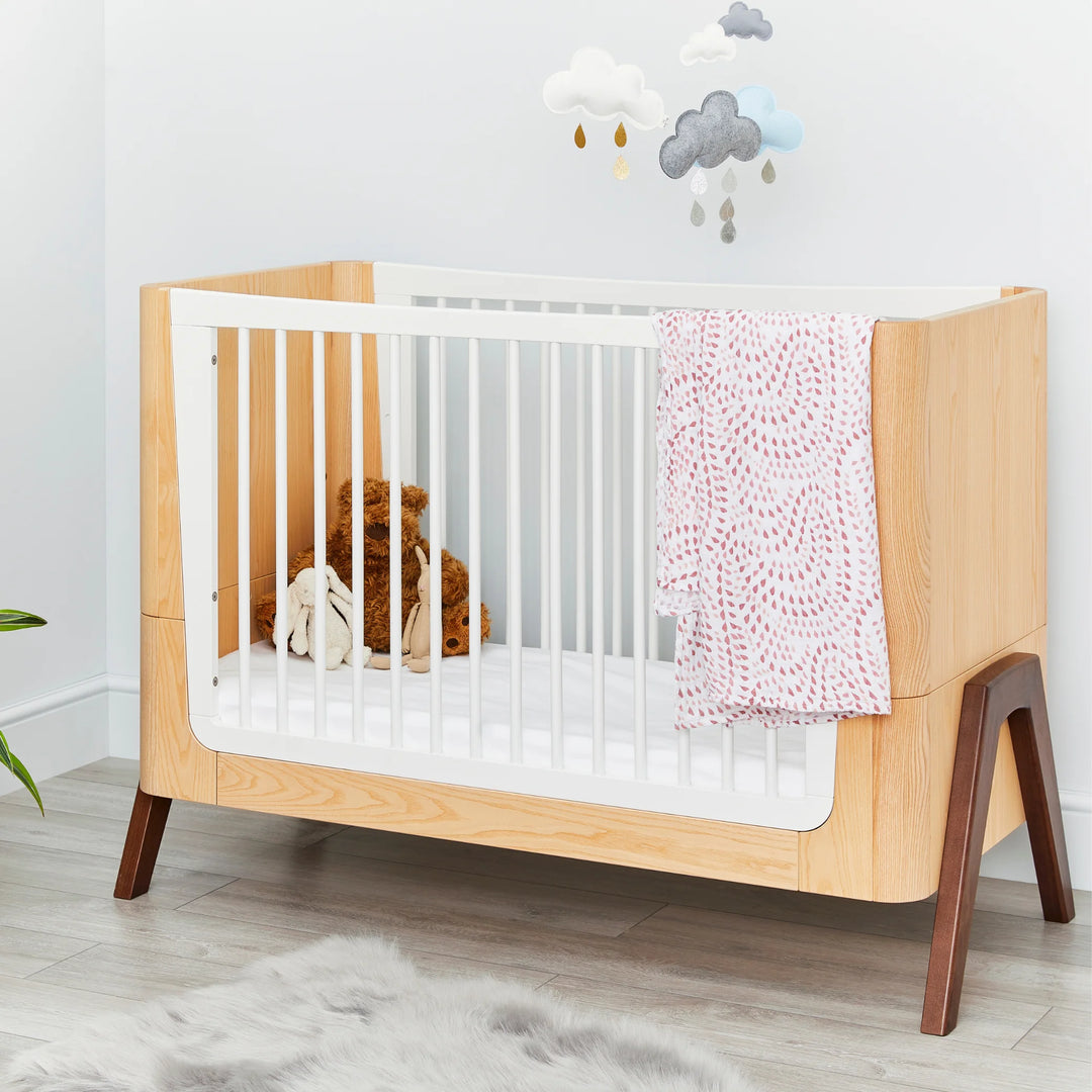 Lifestyle nursery scene featuring the Gaia Baby Hera cot in ash and walnut with white slatted sides, styled with soft blankets, plush toys and cloud wall décor — premium Scandinavian sustainable baby furniture in a modern room.