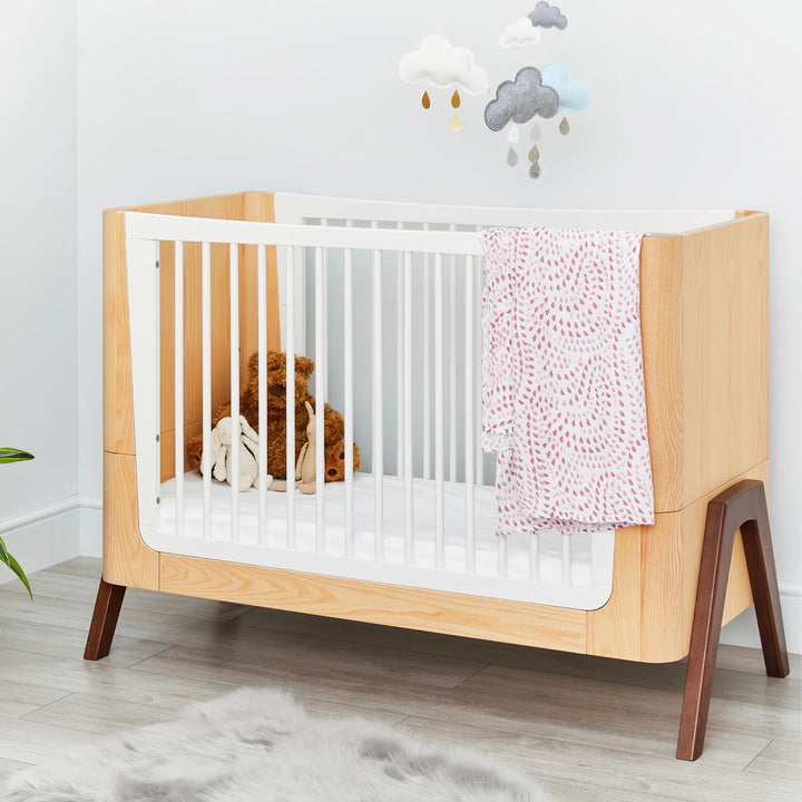 Lifestyle nursery scene featuring the Gaia Baby Hera cot in ash and walnut with white slatted sides, styled with soft blankets, plush toys and cloud wall décor — premium Scandinavian sustainable baby furniture in a modern room.