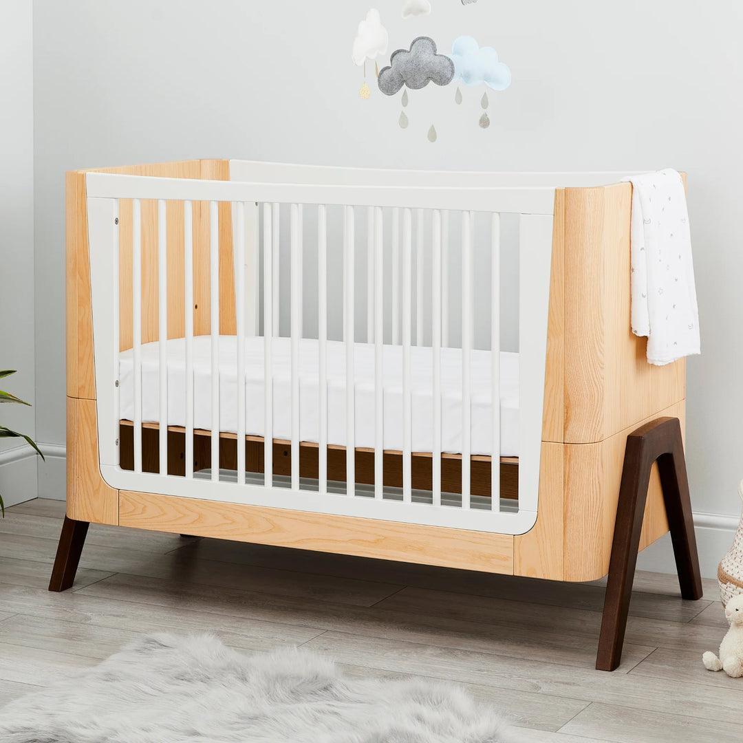 Lifestyle nursery featuring the Gaia Baby Hera cot in ash and walnut with white slatted sides, natural wood panels and solid walnut A-frame legs, styled with soft bedding and cloud wall décor — premium Scandinavian sustainable baby furniture.