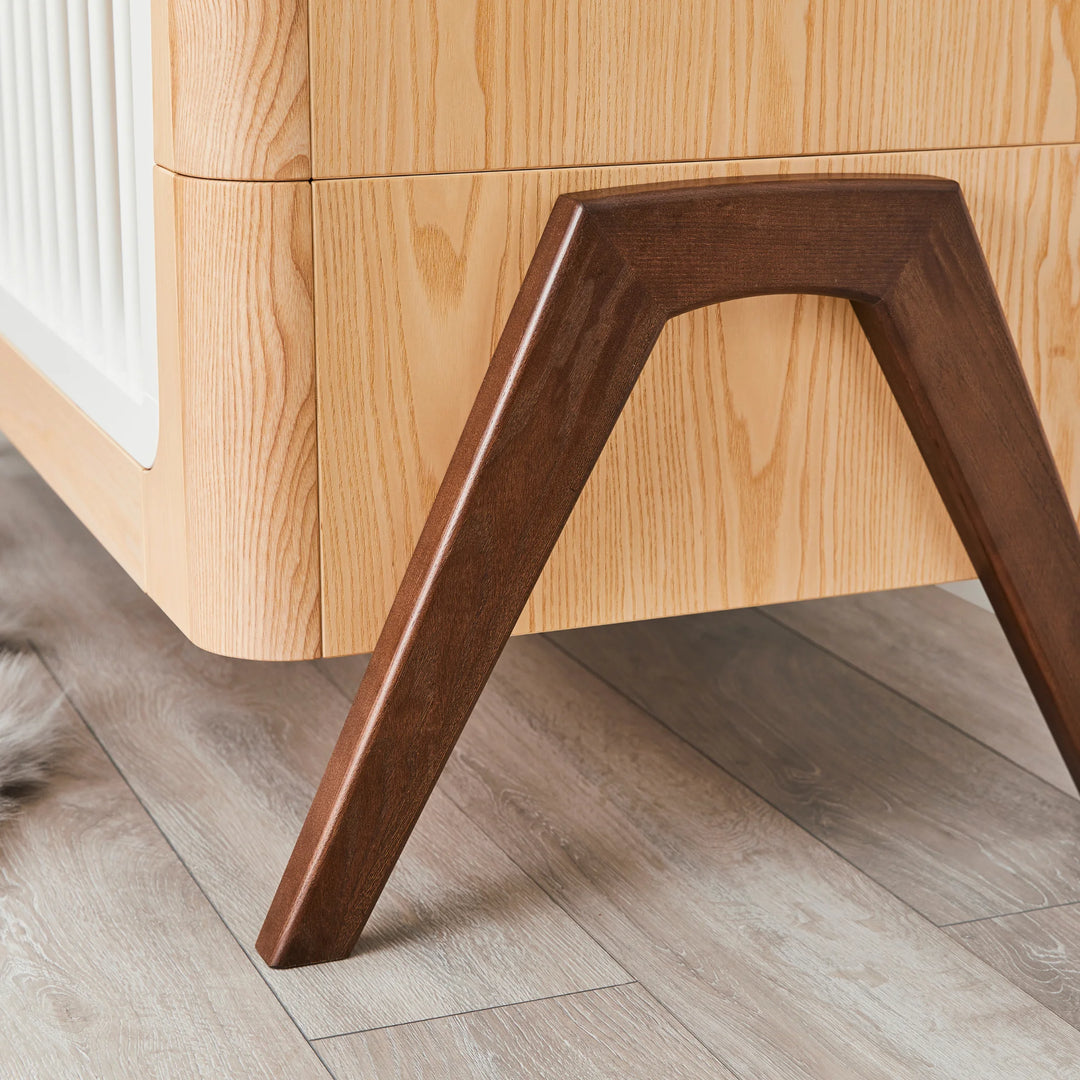 Close-up of the Gaia Baby Hera cot in ash and walnut, showing solid walnut A-frame legs, natural ash wood grain and curved Scandinavian design — premium sustainable nursery furniture craftsmanship.