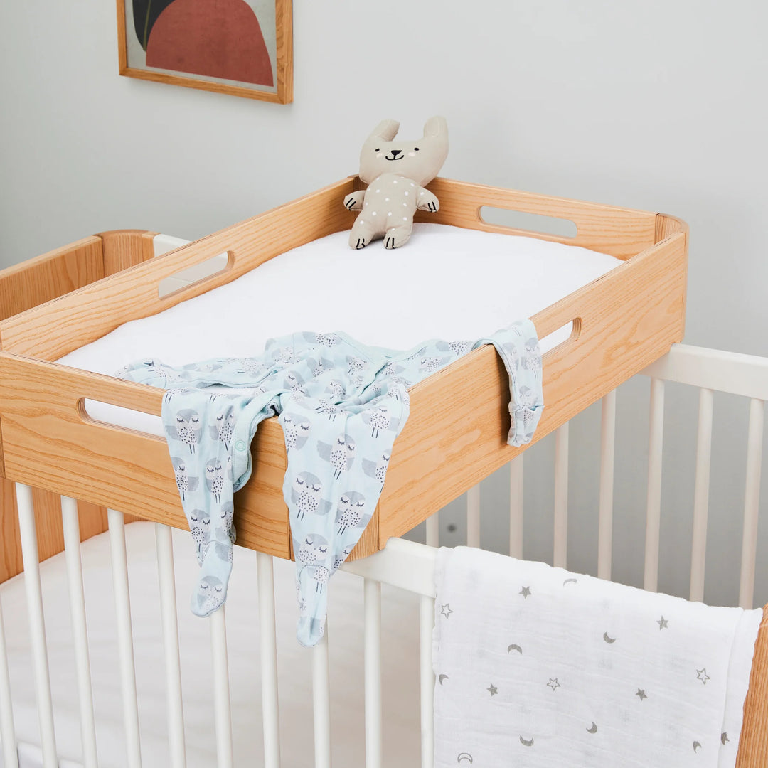 Gaia Baby Hera cot in ash and walnut with matching wooden changing tray on top, styled with soft baby clothes and plush toy — premium Scandinavian sustainable nursery furniture and accessories.