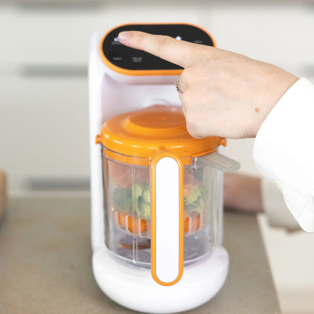 Parent using Quook by Quark 5-in-1 baby food maker — pressing digital control panel to steam fresh vegetables.