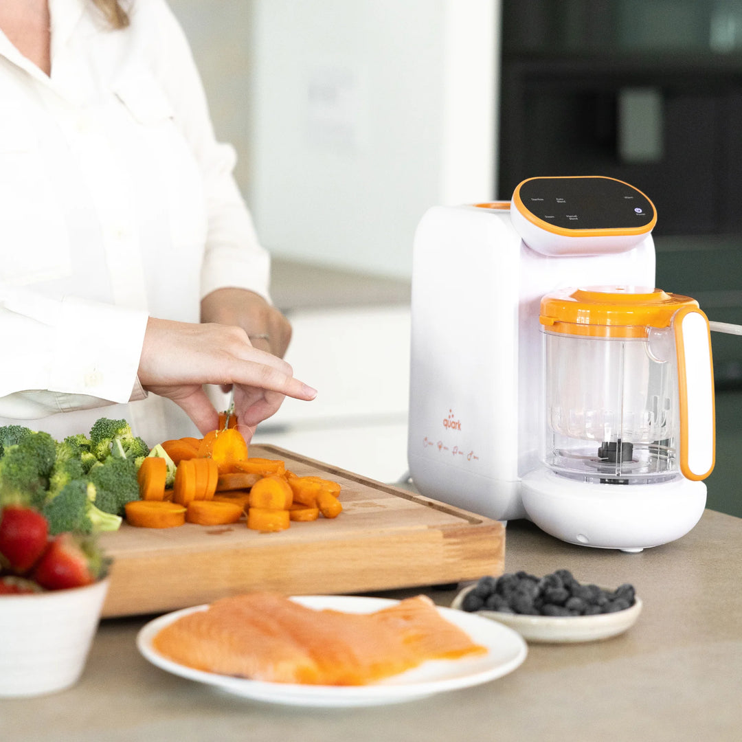 Parent preparing fresh vegetables beside Quook multifunctional steamer, blender, and steriliser for healthy homemade baby meals in modern kitchen setting.