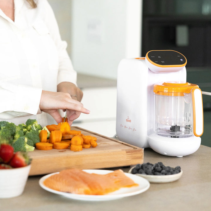 Parent preparing fresh vegetables beside Quook multifunctional steamer, blender, and steriliser for healthy homemade baby meals in modern kitchen setting.