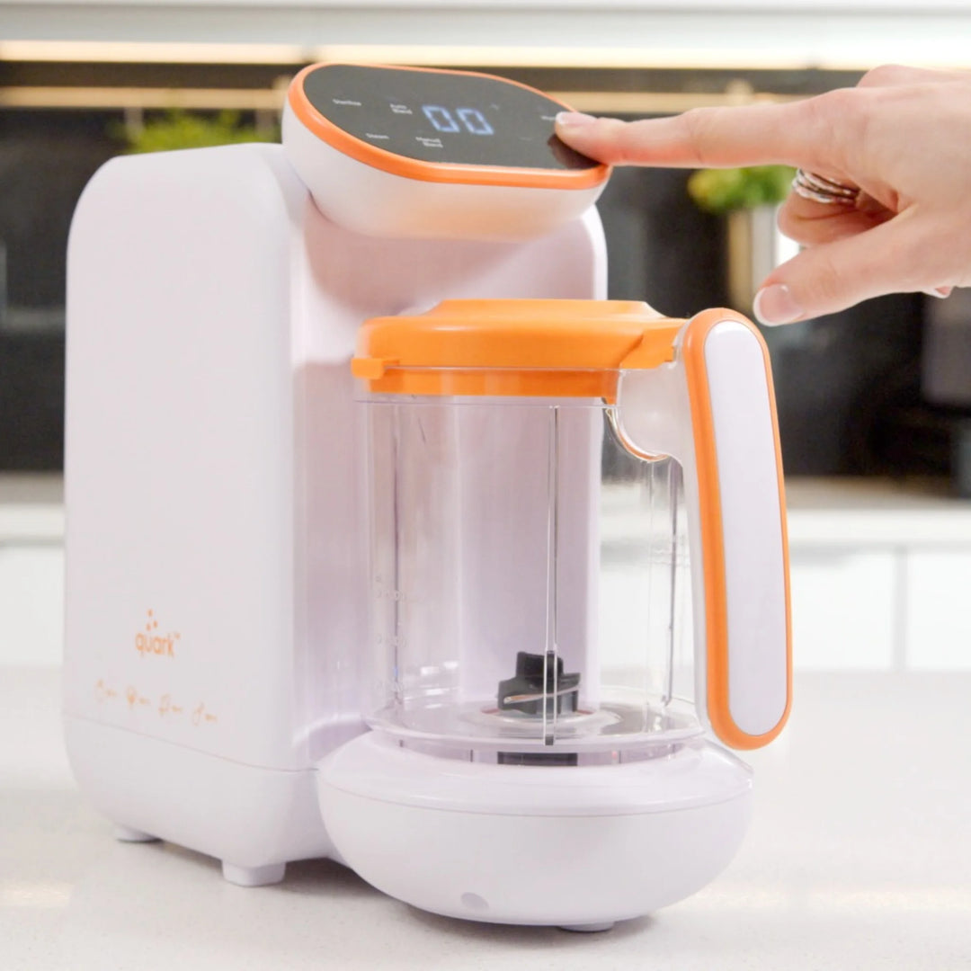 Parent pressing digital touchscreen on Quook by Quark 5-in-1 baby food maker — smart multifunctional baby food processor for steaming, blending, warming and sterilising homemade baby meals in sleek white and orange design.