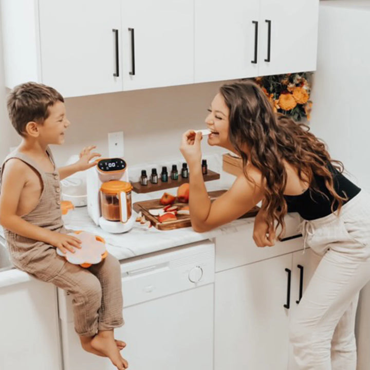 Mother and child preparing healthy snacks together using Quook 5-in-1 baby food maker, multifunctional steamer and blender for homemade baby meals, shown in bright modern kitchen with fresh fruit.