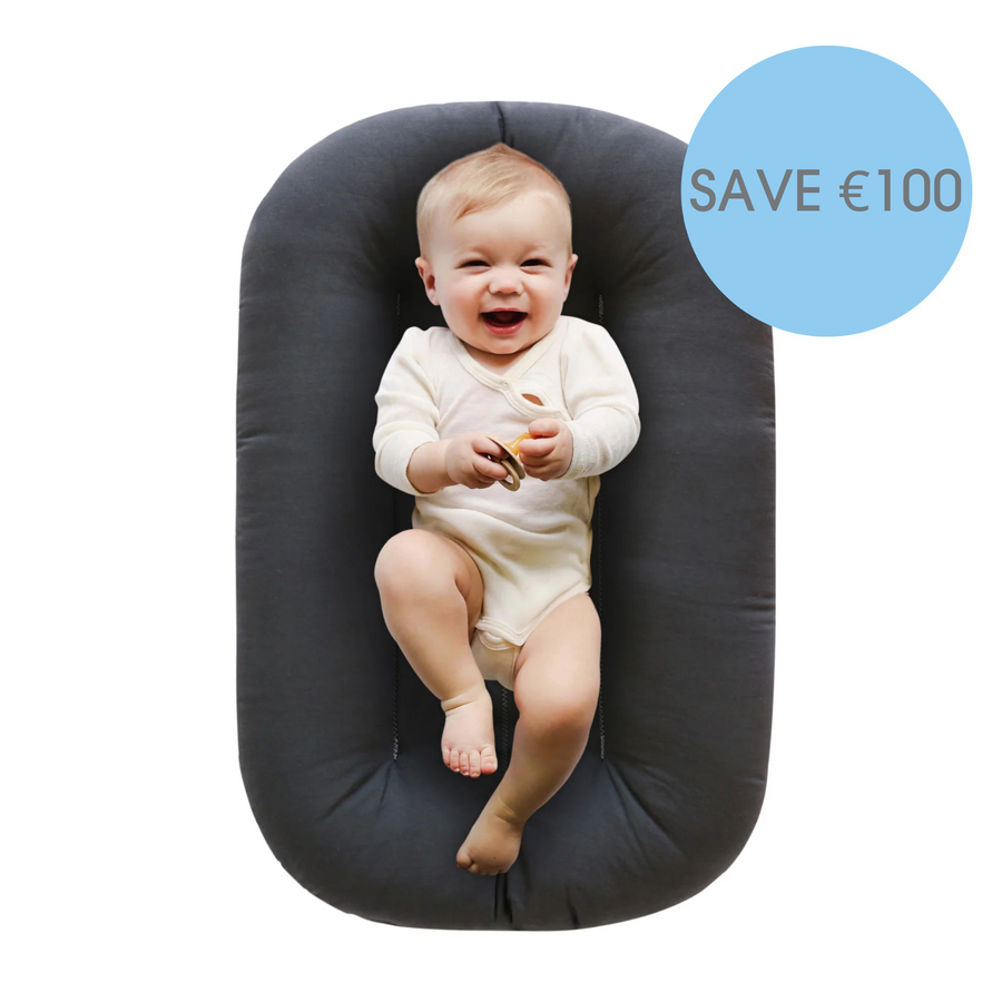 SnuggleMe Newborn infant lounger in sparrow with a cute baby lying with a soother in their hands and smiling at the camera. There is a blue bubble in the top right of this picture showing the saving of €100