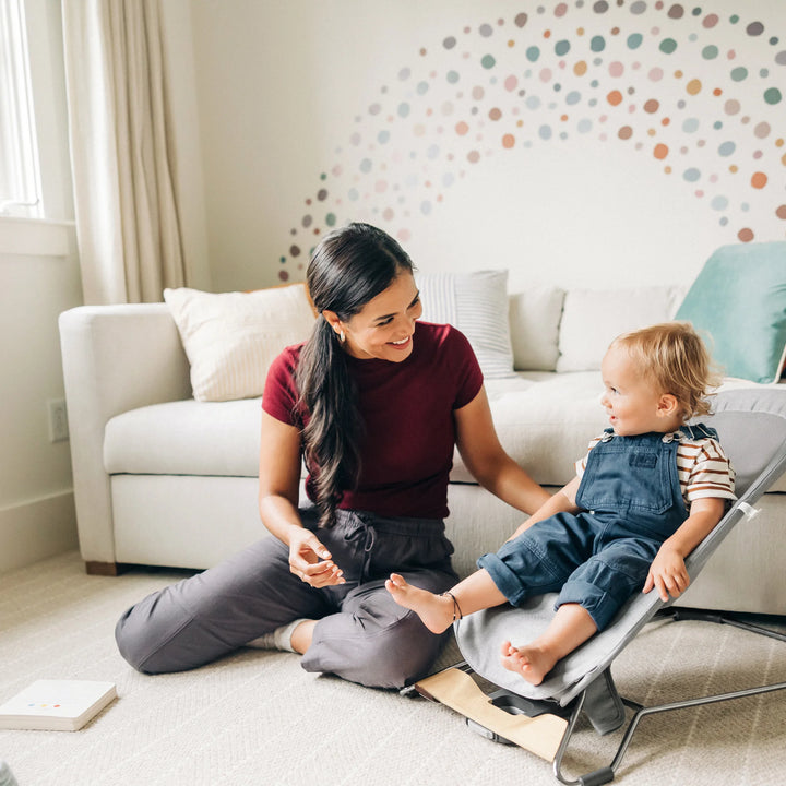 Toddler sitting in the UPPAbaby Mira Bouncer in Stella grey while a parent plays nearby, showcasing the ergonomic toddler-support seat, breathable mesh design, and modern lightweight frame with wood-accented base in a bright family living room.