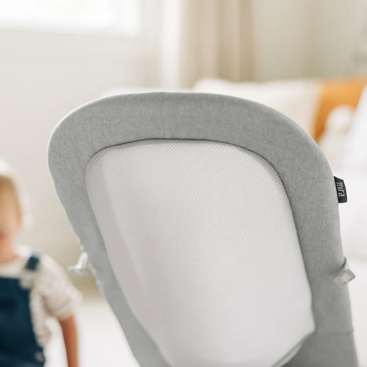 Close-up of the UPPAbaby Mira Bouncer in Stella grey showcasing its breathable mesh back panel and soft textured fabric, photographed in a bright family living room with a child in the background.