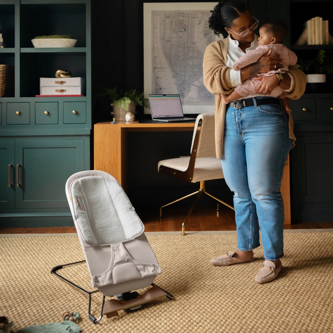 Parent holding baby in a modern home office with the UPPAbaby Mira Bouncer in Charlie beige positioned nearby, showcasing the ergonomic newborn seat, breathable mesh design, and stylish wooden-base frame for everyday family use.