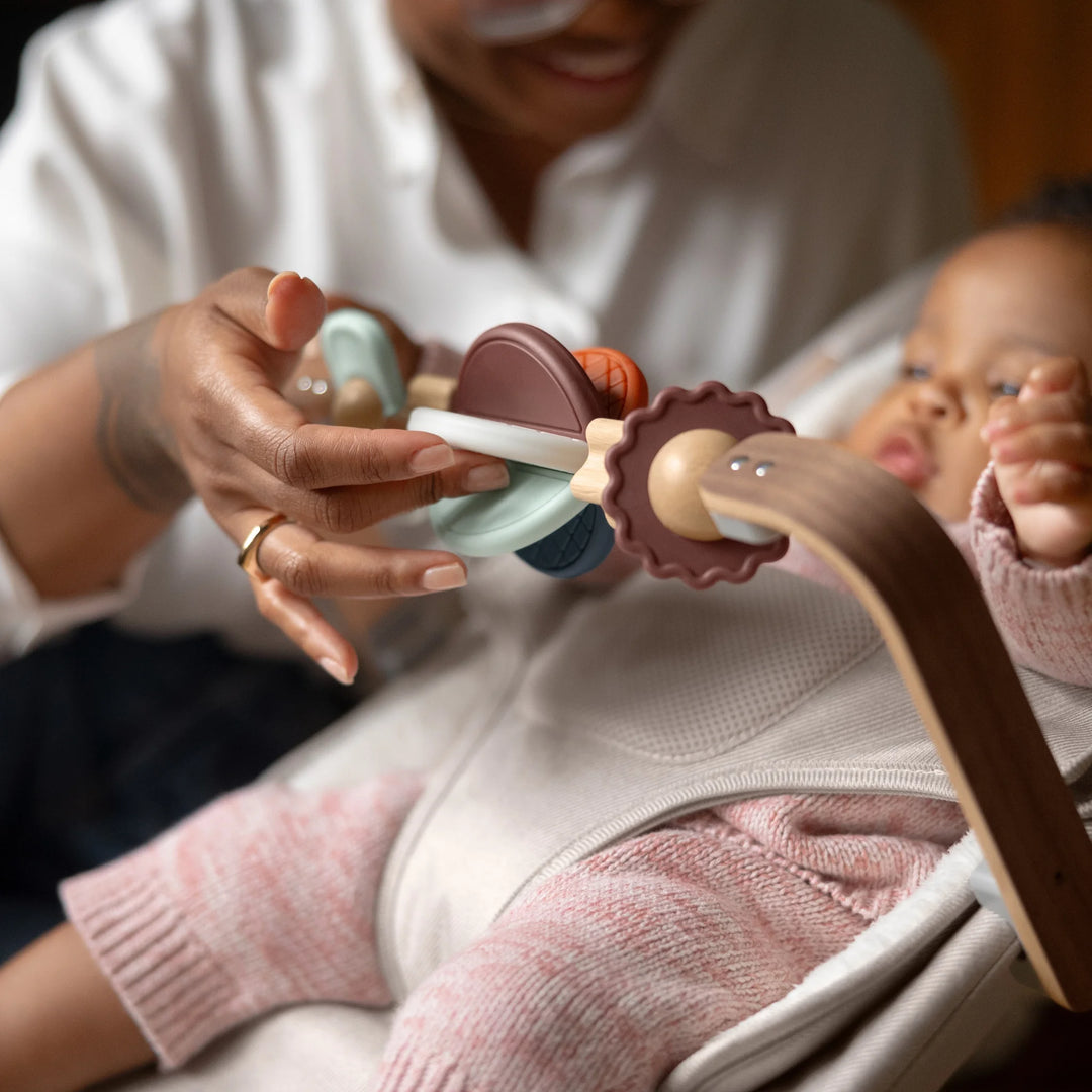 Parent engaging baby with the UPPAbaby Mira Bouncer wooden toy bar accessory while the baby sits securely in the Mira Bouncer in Charlie beige, highlighting developmental play features and ergonomic infant support.”