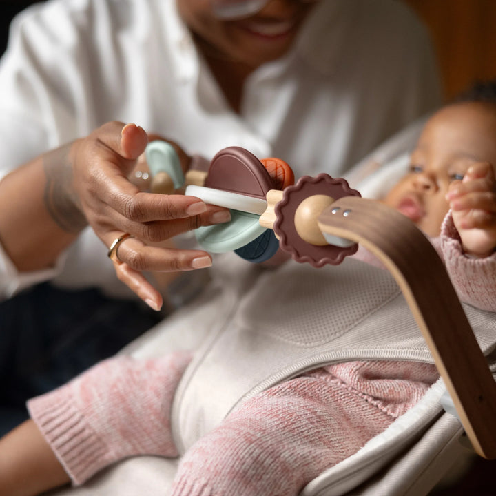 Parent engaging baby with the UPPAbaby Mira Bouncer wooden toy bar accessory while the baby sits securely in the Mira Bouncer in Charlie beige, highlighting developmental play features and ergonomic infant support.”
