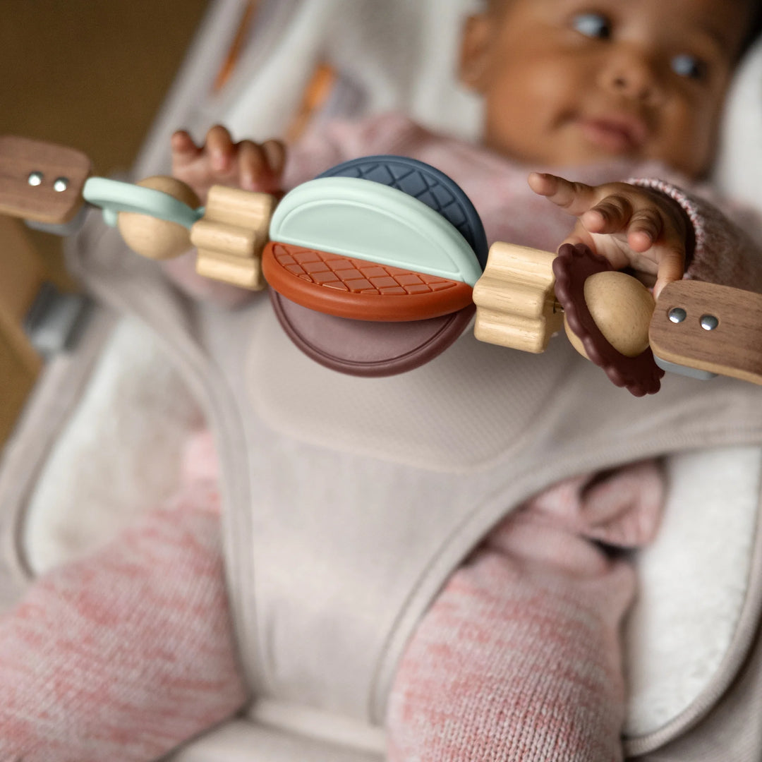 Baby playing with the UPPAbaby Mira Bouncer wooden toy bar accessory while seated in the Mira Bouncer in Charlie beige, showing colourful developmental toys and secure ergonomic harness in a cozy home setting.”