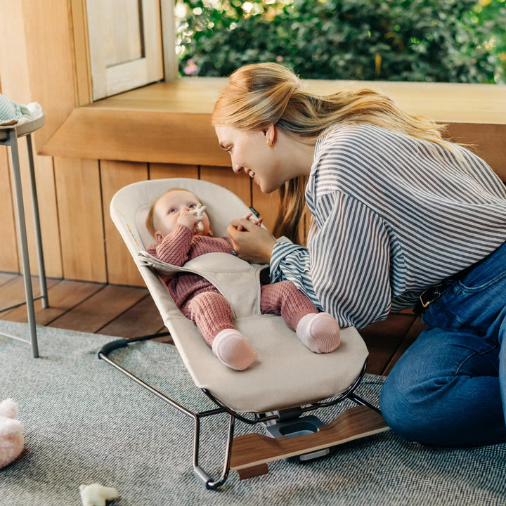 “Mother interacting with her baby seated in the UPPAbaby Mira Bouncer in Charlie beige, showing ergonomic newborn support, breathable mesh design, and modern wooden-base frame in a bright, cozy home setting.”