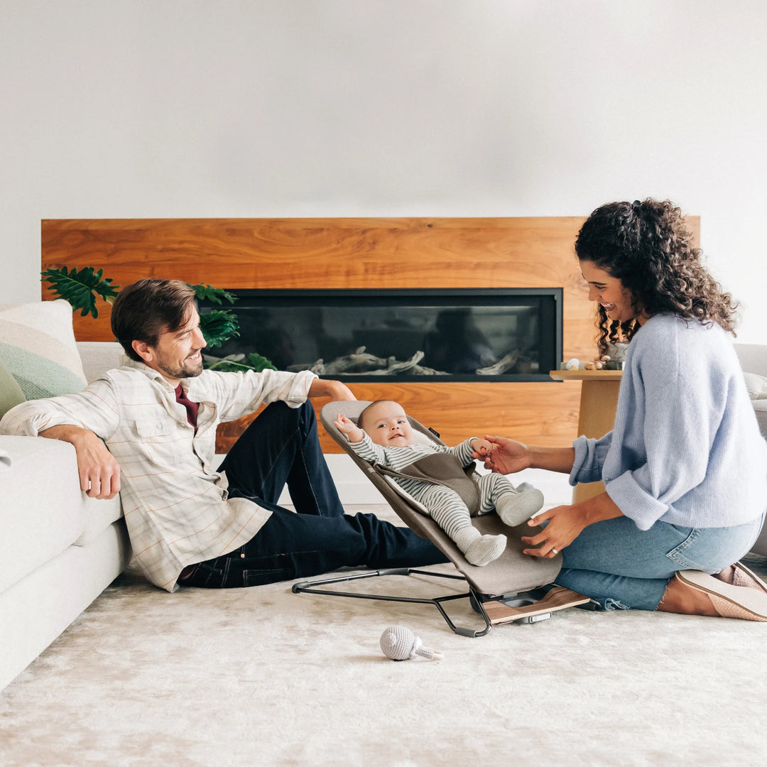 “Parents interacting with baby seated in UPPAbaby Mira Bouncer in Wells fabric, shown in a modern living room with soft neutral décor, ergonomic baby seat design, and supportive newborn insert—premium baby bouncer for comfort, bonding, and developmental play.”