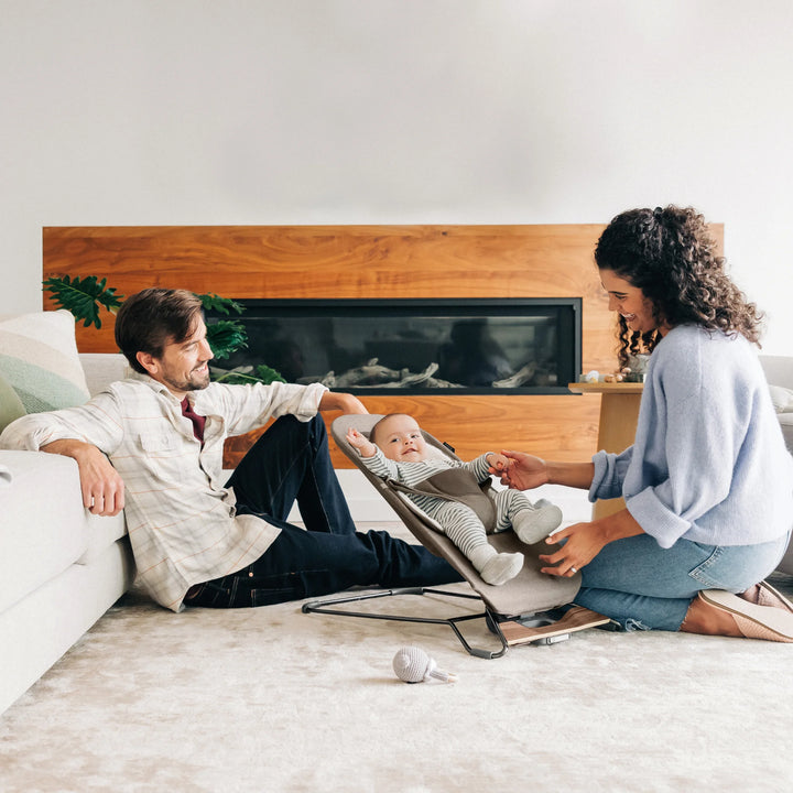 “Parents interacting with baby seated in UPPAbaby Mira Bouncer in Wells fabric, shown in a modern living room with soft neutral décor, ergonomic baby seat design, and supportive newborn insert—premium baby bouncer for comfort, bonding, and developmental play.”