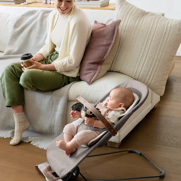 “Mother relaxing on sofa while baby plays in UPPAbaby Mira Bouncer in Wells fabric, featuring wooden toy bar, ergonomic infant support, and modern living room setting—premium baby bouncer for comfort, play, and development.”
