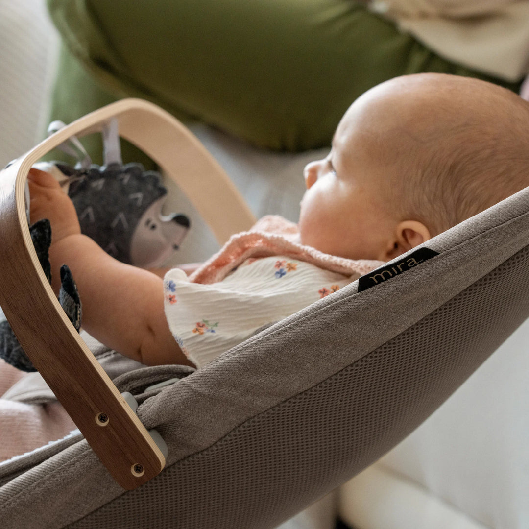 “Baby relaxing in the UPPAbaby Mira Bouncer in Wells with supportive mesh seat and natural wood toy bar, engaging with soft plush animal toys in a cozy home setting.”