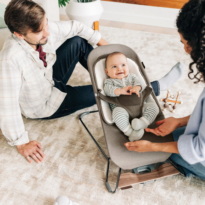 Parents engaging with their baby seated in the UPPAbaby Mira Bouncer in Wells taupe, highlighting the ergonomic infant support, plush newborn insert, breathable mesh design, and modern wood-accented frame in a bright family living room.