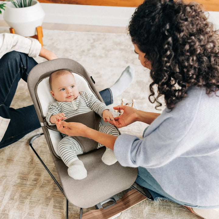 Parents securing their baby in the UPPAbaby Mira Bouncer in Wells taupe, highlighting the ergonomic newborn support, breathable mesh design, and modern wooden-base frame in a bright family living room.