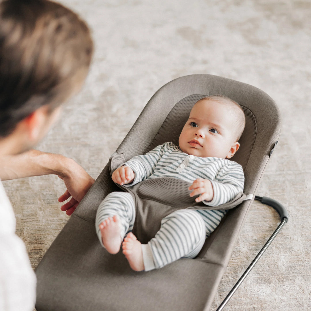Baby relaxing in the UPPAbaby Mira Bouncer in Wells taupe while interacting with a parent, showcasing the ergonomic infant seat, breathable mesh support, and modern minimalist frame in a bright family living space.