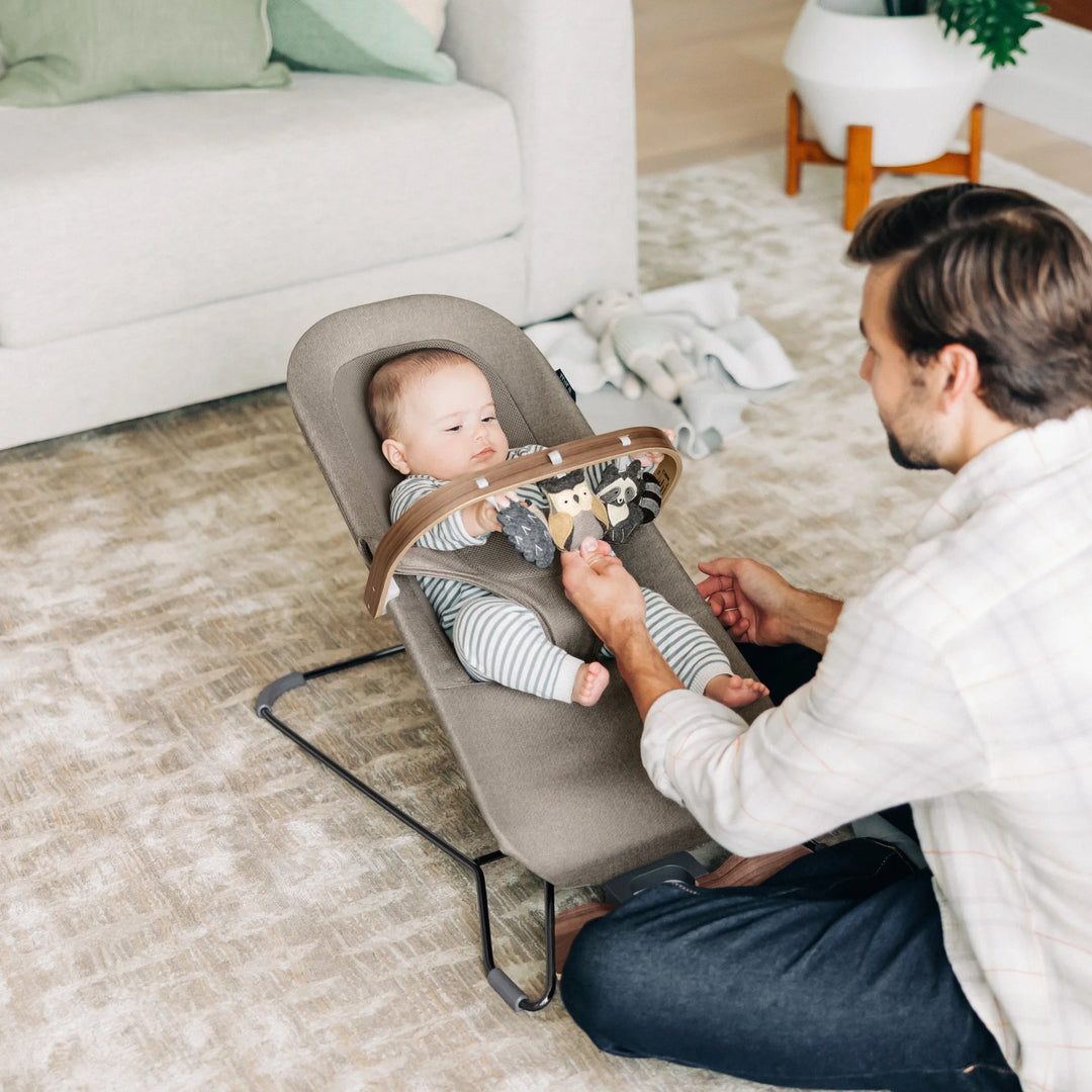 “Father playing with baby in UPPAbaby Mira Bouncer in Wells fabric, using wooden toy bar in modern living room—premium ergonomic baby bouncer for interactive play, comfort, and infant development.”