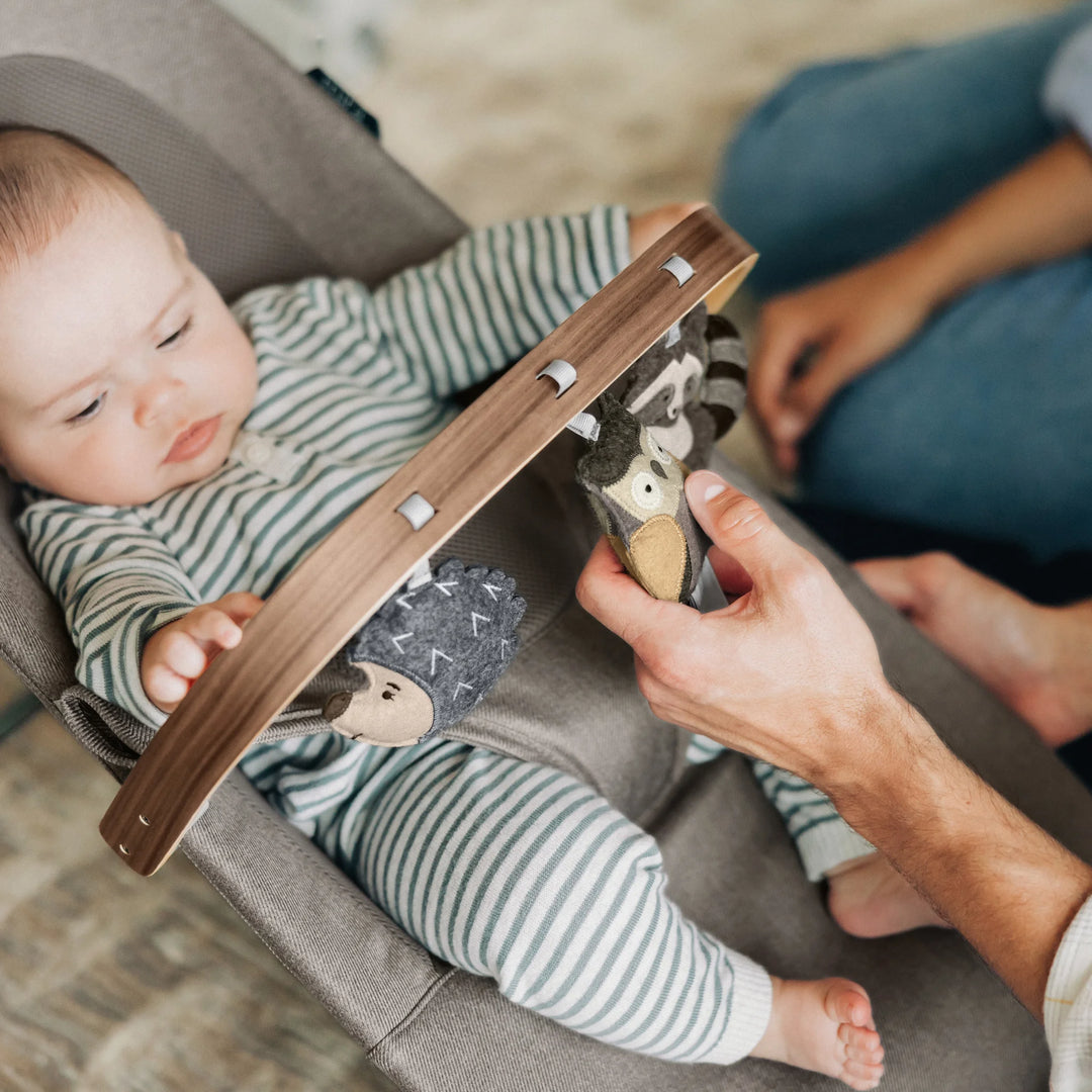 “Baby playing with wooden toy bar on UPPAbaby Mira Bouncer in Wells fabric, engaging with sensory plush characters while parent assists—premium ergonomic baby bouncer for interactive playtime.”