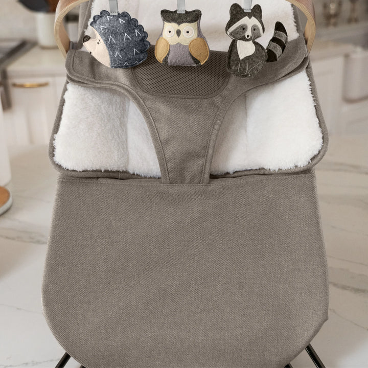 “UPPAbaby Mira Bouncer in Wells taupe with plush newborn insert and wooden toy bar featuring soft animal toys—hedgehog, owl, and raccoon—shown in a modern kitchen setting for enhanced infant entertainment and comfort.”