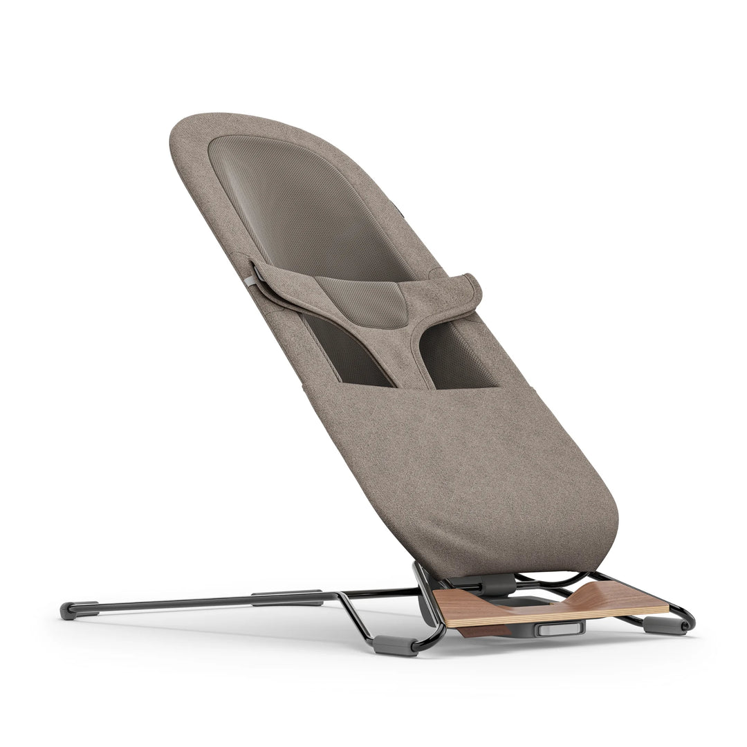 “UPPAbaby Mira Bouncer in Wells taupe shown in upright angled view, featuring breathable mesh seat panel, ergonomic infant harness, textured fabric upholstery, and sleek lightweight frame with walnut-tone wood base accent.”