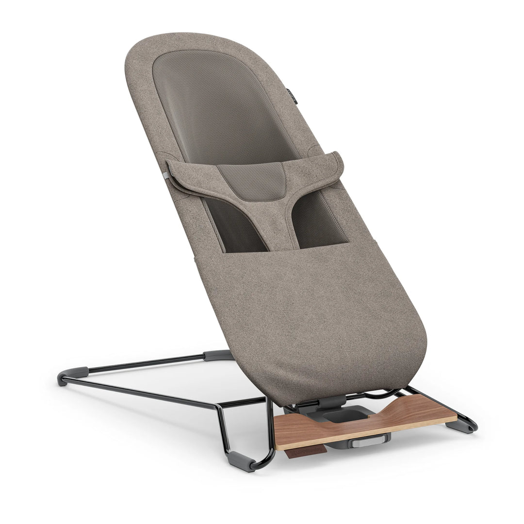 UPPAbaby Mira Bouncer in Wells taupe shown in angled upright view, featuring breathable mesh seat panel, ergonomic infant harness, textured taupe upholstery, and modern lightweight steel frame with walnut-tone wood base accent.