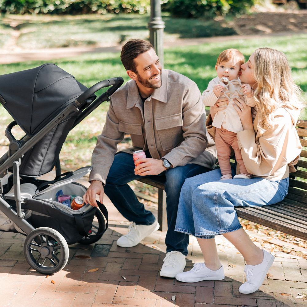 The Bevvy stroller basket cooler from UPPAbaby is designed to seamlessly fit in the basket of the Vista, Cruz and Ridge models. This image depicts a family with their child sitting on a bench in a sunny park with the dad taking out a fizzy drink can from the basket cooler.