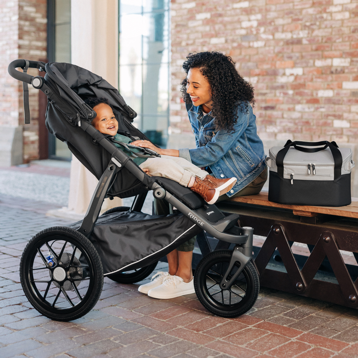 A mother is smiling widely at her child in the Ridge jogging stroller from UPPAbaby on a sheltered place on a sunny day. The Bevvy stroller basket cooler has been taken out of the basket and is sitting next to the Mum on the bench,