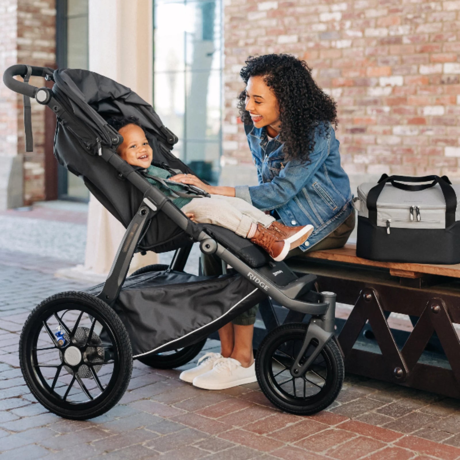 UPPAbaby Bevvy stroller basket cooler bag shown beside UPPAbaby pushchair during family outing, insulated cooler for drinks and snacks on the go.