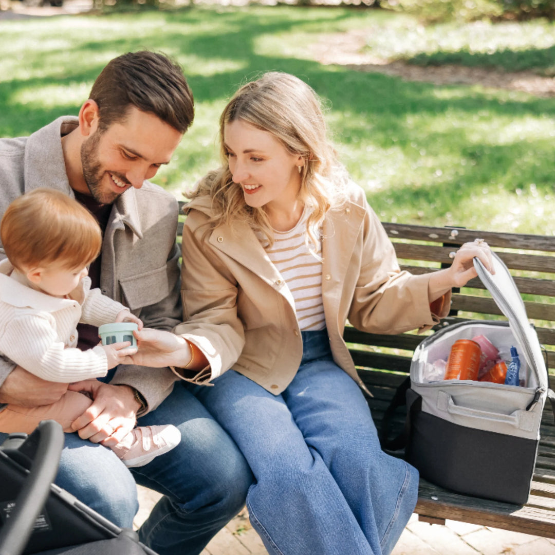 UPPAbaby Bevvy stroller basket cooler bag opened on park bench during family outing, insulated cooler holding drinks and snacks for use with UPPAbaby pushchairs.